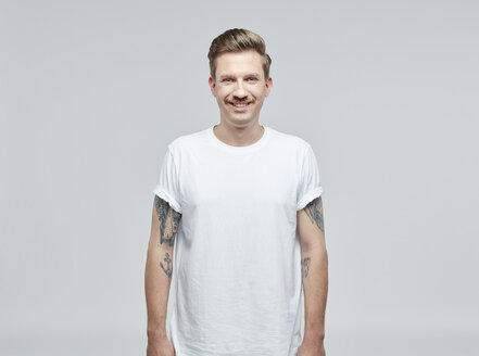 Porträt eines lächelnden Mannes mit Tatoos auf den Armen, der ein weißes T-Shirt vor einem grauen Hintergrund trägt - RHF001090