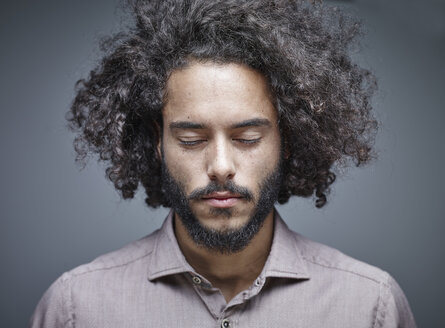 Porträt eines bärtigen jungen Mannes mit lockigem braunem Haar und geschlossenen Augen vor grauem Hintergrund - RHF001100