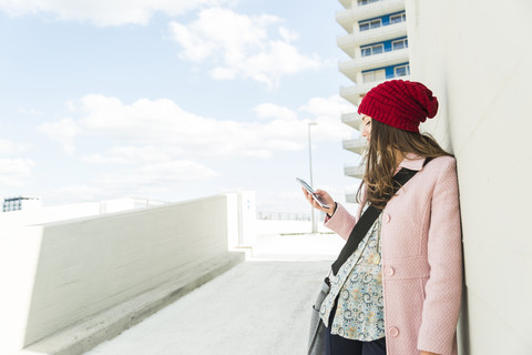 Junge Frau lehnt an einer Betonwand und schaut auf ihr Handy, lizenzfreies Stockfoto