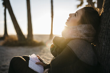 Spain, Tarragona, pensive young woman with notebook sitting on the beach at twilight - JRFF000234