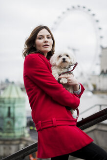 UK, London, Porträt einer jungen Frau mit Hund auf dem Arm und London Eye im Hintergrund - MAUF000156