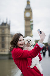 Großbritannien, London, Porträt einer lächelnden jungen Frau mit Hund auf dem Arm, die ein Selfie mit ihrem Smartphone macht - MAUF000158