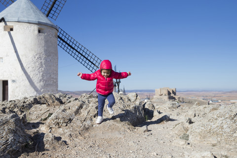 Spanien, Consuegra, springendes kleines Mädchen mit Windmühle im Hintergrund, lizenzfreies Stockfoto