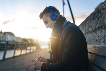 Irland, Dublin, junger Mann sitzt auf einer Bank im Gegenlicht und hört Musik mit Kopfhörern - BOYF000085
