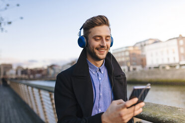Irland, Dublin, junger Mann hält Smartphone und hört Musik mit Kopfhörern - BOYF000087