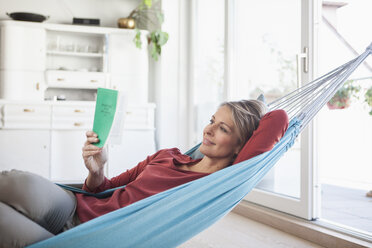 Smiling woman at home lying in hammock reading book - RBF003569