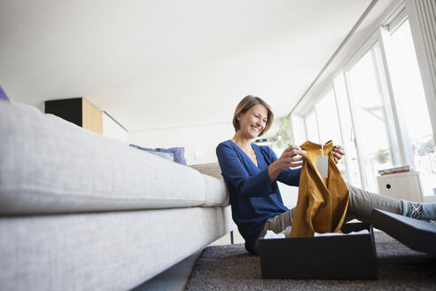 Smiling woman at home unpacking parcel with garment - RBF003593