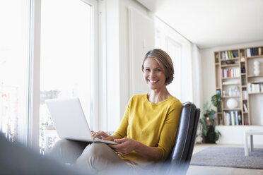 Glückliche Frau zu Hause auf einem Ledersessel sitzend mit einem Laptop - RBF003602