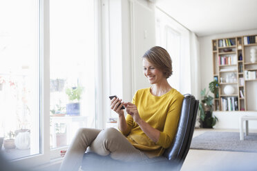 Entspannte Frau zu Hause, die auf einem Ledersessel sitzt und ein Mobiltelefon benutzt - RBF003603