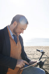 Spanien, Barcelona, lächelnder Geschäftsmann mit Fahrrad und digitalem Tablet vor dem Strand - VABF000005