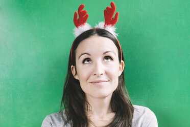 Portrait of smiling woman wearing hair-band with red reindeer antlers in front of green wall - GEMF000546