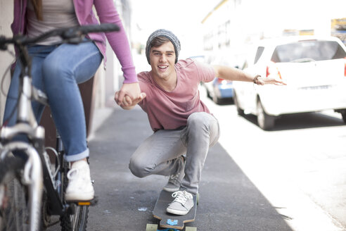 Teenage girl on bicycle pushing young man on skateboard - ZEF007593