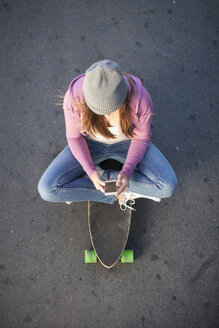 Teenage girl sitting on skateboard looking at cell phone - ZEF007615