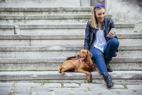 Lächelnde Frau mit Smartphone sitzt neben ihrem Hund auf der Treppe - ZEDF000030