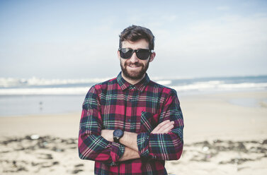 Spain, La Coruna, portrait of smiling hipster with sunglasses standing on the beach - RAEF000726