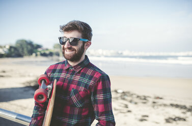 Spain, La Coruna, portrait of smiling hipster wearing sunglasses with his skateboard on the beach - RAEF000727