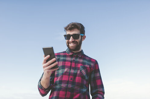 Spanien, La Coruna, Porträt eines lächelnden Hipsters mit Sonnenbrille, der vor blauem Himmel auf sein Phablet schaut - RAEF000728