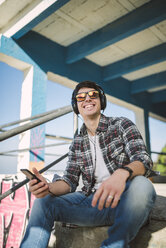 Porträt eines lächelnden jungen Mannes mit verspiegelter Sonnenbrille, der mit seinen Kopfhörern auf einer Treppe sitzt - RAEF000729