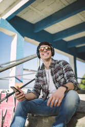 Portrait of smiling young man with mirrored sunglasses sitting on stairs with his headphones - RAEF000729