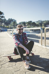 Spanien, La Coruna, Porträt eines lächelnden Hipsters mit Sonnenbrille, der auf seinem Longboard sitzt - RAEF000734