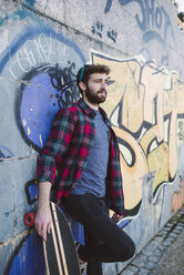Spain, La Coruna, portrait of hipster leaning against wall with his longboard - RAEF000735