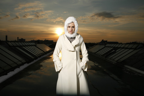 Porträt einer Frau mit weißem Mantel und Kapuze auf einem Dach in der Abenddämmerung, lizenzfreies Stockfoto