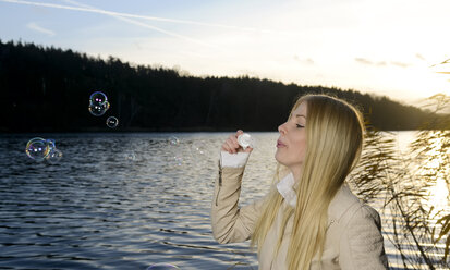 Blond young woman blowing soap bubbles in front of a lake - BFRF001709