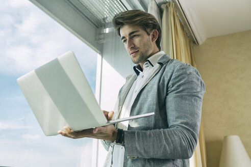 Porträt eines jungen Geschäftsmannes, der vor einem Fenster steht und einen Laptop hält - TAMF000349