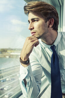 Porträt eines jungen Geschäftsmannes mit Hand am Kinn, der durch ein Fenster schaut - TAM000350