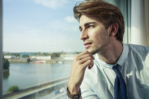 Porträt eines jungen Geschäftsmannes mit Hand am Kinn, der durch ein Fenster schaut - TAM000351