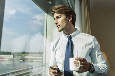 Portrait of young businessman with smartphone and mug looking through window - TAM000352