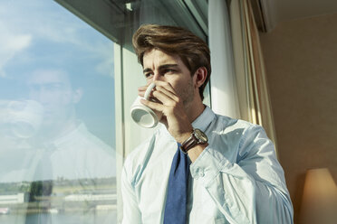 Young businessman with mug looking through window - TAM000353