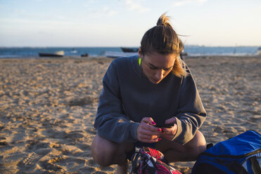 Spain, Puerto Real, young woman looking at her smartphone while crouching on the beach - KIJF000052