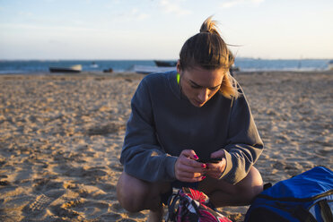 Spanien, Puerto Real, junge Frau schaut auf ihr Smartphone, während sie am Strand hockt - KIJF000052