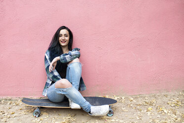 Porträt einer lächelnden jungen Frau, die auf einem Longboard vor einer rosa Wand sitzt - KIJF000053