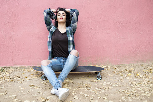 Porträt einer jungen Frau, die auf einem Longboard sitzt und sich vor einer rosa Wand entspannt - KIJF000054