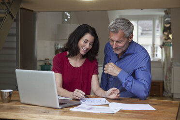 Lächelndes Paar zu Hause mit Laptop und Papieren am Tisch - RBF003645