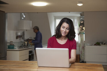 Smiling woman at home using laptop with man in background - RBF003648