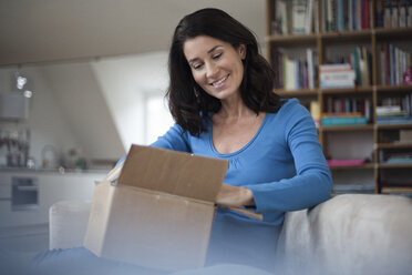 Smiling woman at home sitting on couch unpacking parcel - RBF003659