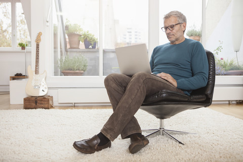 Älterer Mann sitzt zu Hause auf einem Stuhl und benutzt einen Laptop, lizenzfreies Stockfoto