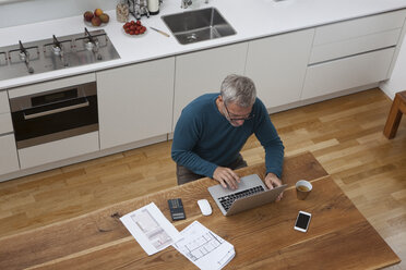 Mature man in kitchen with construction plan using laptop - RBF003721