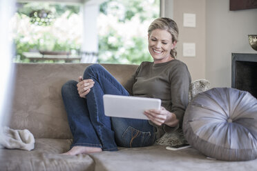 Smiling woman sitting on couch looking at digital tablet - ZEF007684