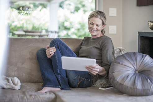 Lächelnde Frau sitzt auf der Couch und schaut auf ein digitales Tablet - ZEF007684
