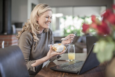 Frau isst Obstsalat und arbeitet zu Hause am Laptop - ZEF007686