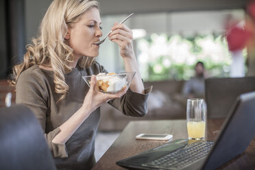 Frau isst Obstsalat und arbeitet zu Hause am Laptop - ZEF007687