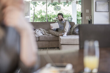 Mann auf Couch, der auf ein digitales Tablet schaut und mit einer Frau im Vordergrund telefoniert - ZEF007688