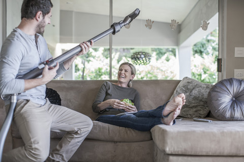 Frau auf Couch und Mann, der so tut, als würde er mit dem Staubsauger Gitarre spielen, lizenzfreies Stockfoto