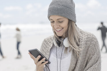Porträt einer lächelnden Frau mit Mütze, die am Strand auf ihr Smartphone schaut - ZEF007714