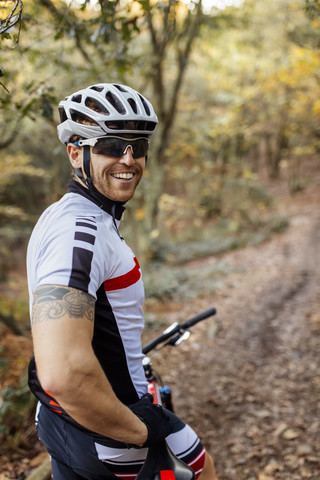Porträt eines lächelnden Mountainbikers mit Fahrradhelm und Sonnenbrille im Wald, lizenzfreies Stockfoto