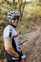 Porträt eines lächelnden Mountainbikers mit Fahrradhelm und Sonnenbrille im Wald - MGOF001216